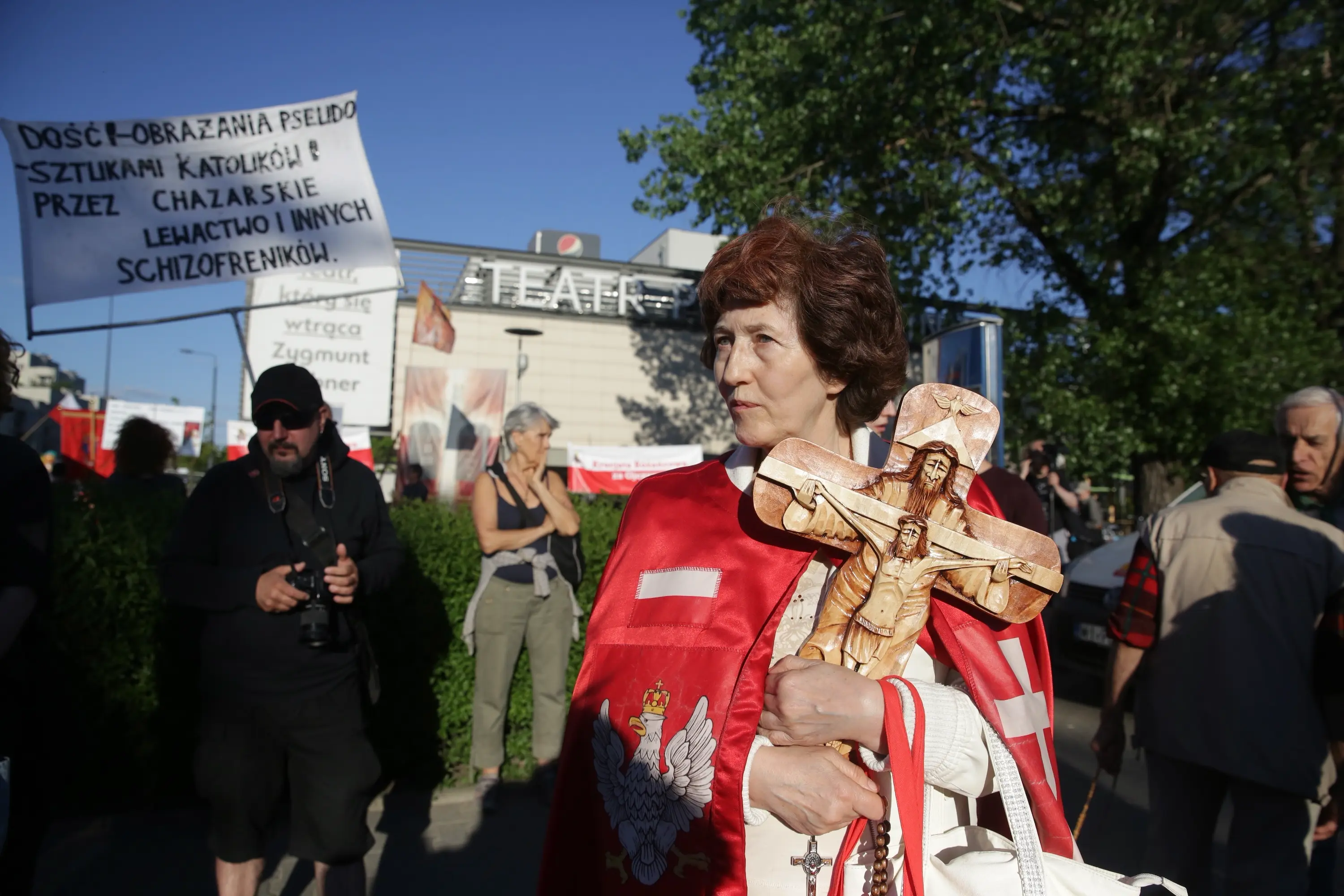  Słoneczny dzień. Grupa protestujących przed Teatrem Powszechnym. Na pierwszym planie starsza kobieta w czerwonej pelerynce z krzyżem i orłem. Do piersi przyciska drewniane figurki ukrzyżowanego Jezusa. W tle transparent „dość obrażania pseudosztukami katolików przez chazarskie lewactwo i innych schizofreników”.