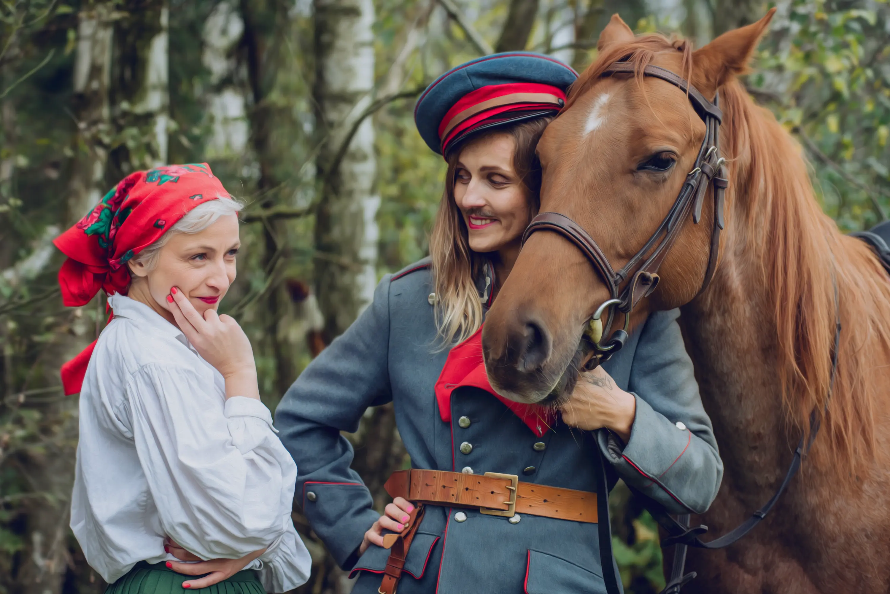 Las. Długowłosa kobieta z wąsami w stroju ułana stoi obok kasztanowego konia. Trzyma lejce. Z uśmiechem spogląda na stojącą obok kobietę. Ta jest w luźnej, białej koszuli i zielonej spódnicy. Białe włosy wystają spod czerwonej, kwiecistej chusty. Z zainteresowaniem obserwuje konia.