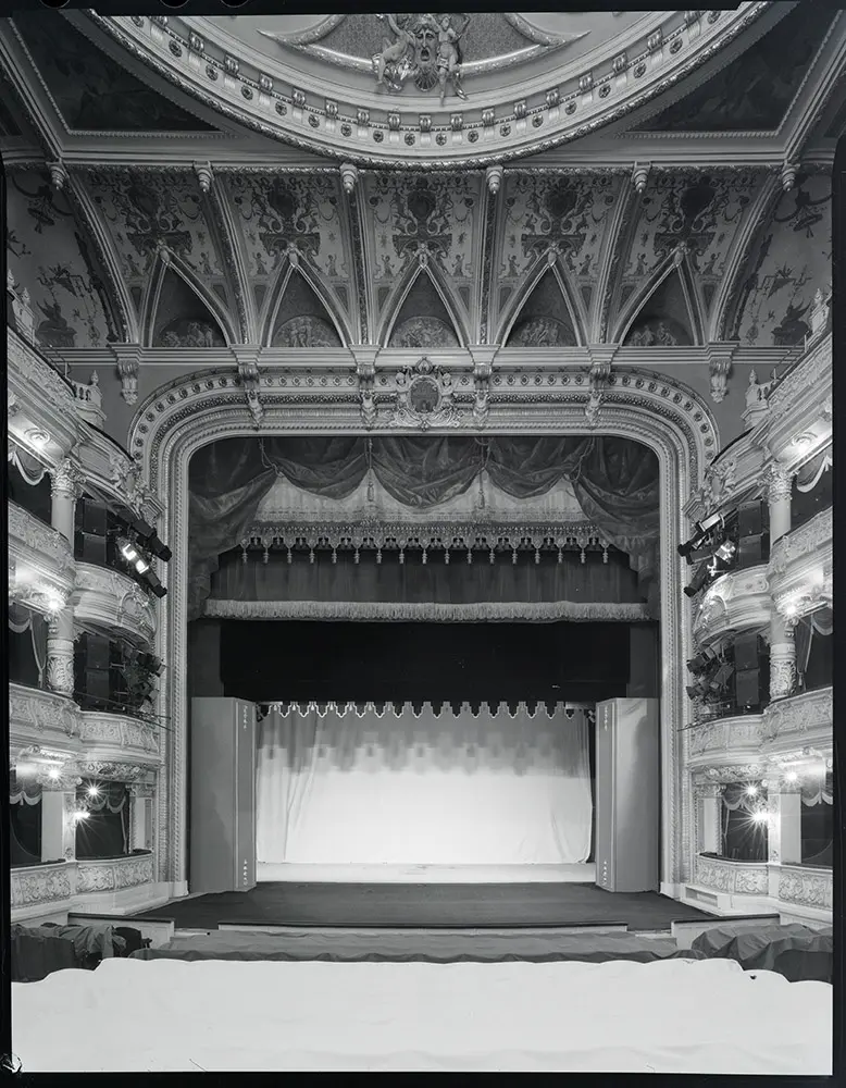 Czarno-białe zdjęcie, widok z widowni Teatru imienia Juliusza Słowackiego w Krakowie. Fotele zakryte ciemnym materiałem. Kurtyna podniesiona. Za nią rozciągnięty biały materiał.