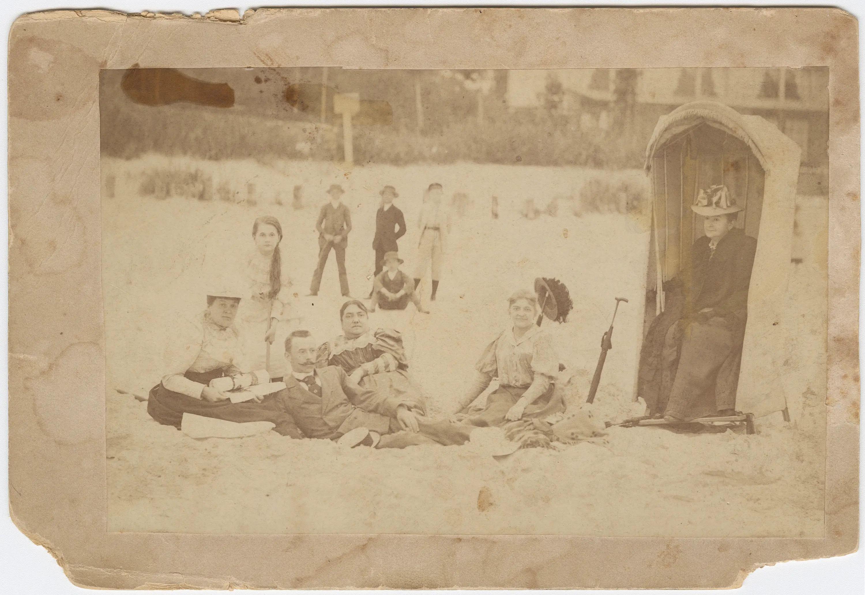  Sepia. Wypłowiałe zdjęcie grupy osób na plaży. Niektórzy siedzą, inni leżą na kocu, Trzy postaci stoją z tyłu, jedna siedzi w plażowym koszu. Wszyscy kompletnie ubrani.