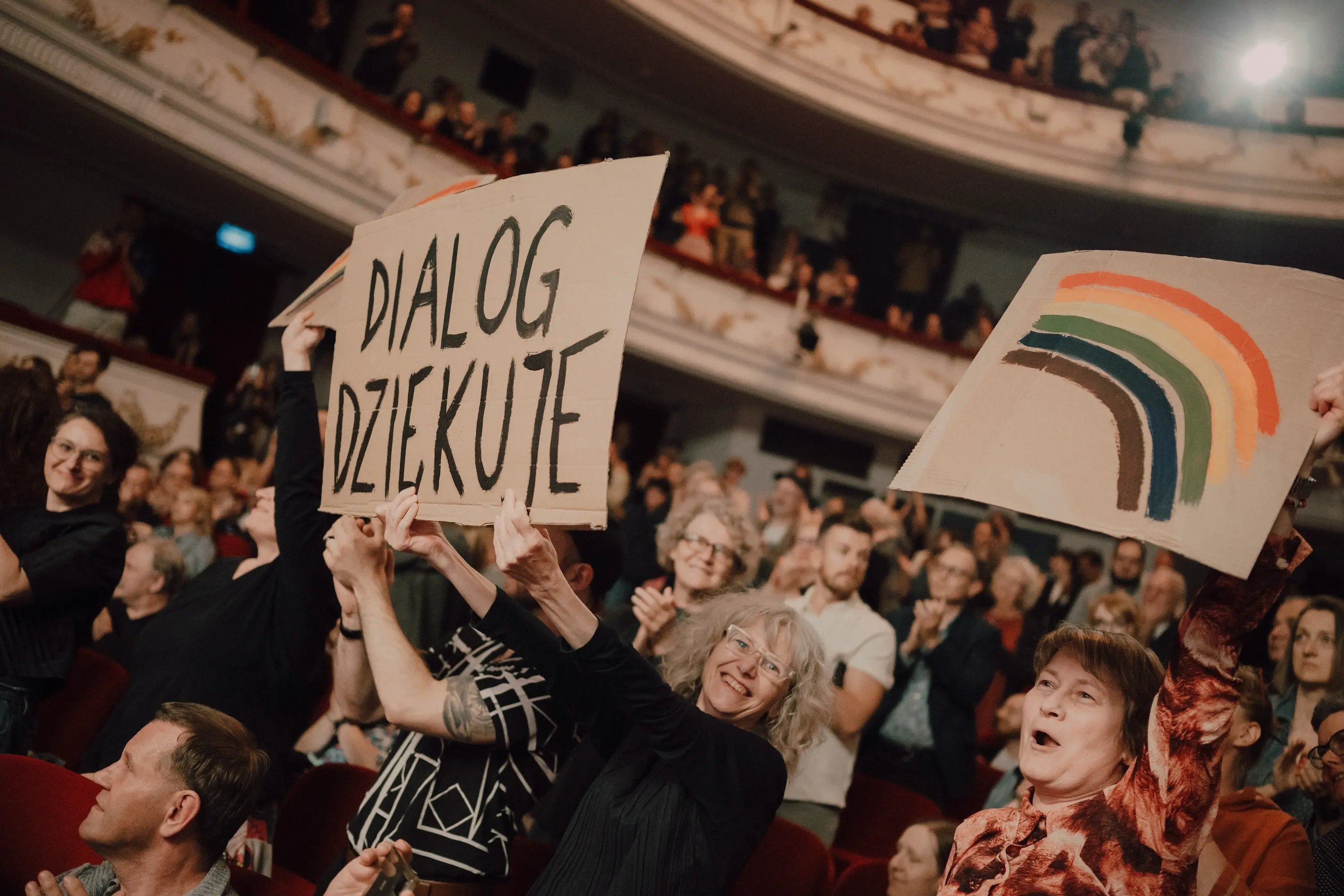 Widownia teatru. Publiczność wstaje z miejsc, klaszcze. Na pierwszym planie kilka osób z transparentami. Na kartonach namalowane tęcze i napis „Dialog dziękuje”.