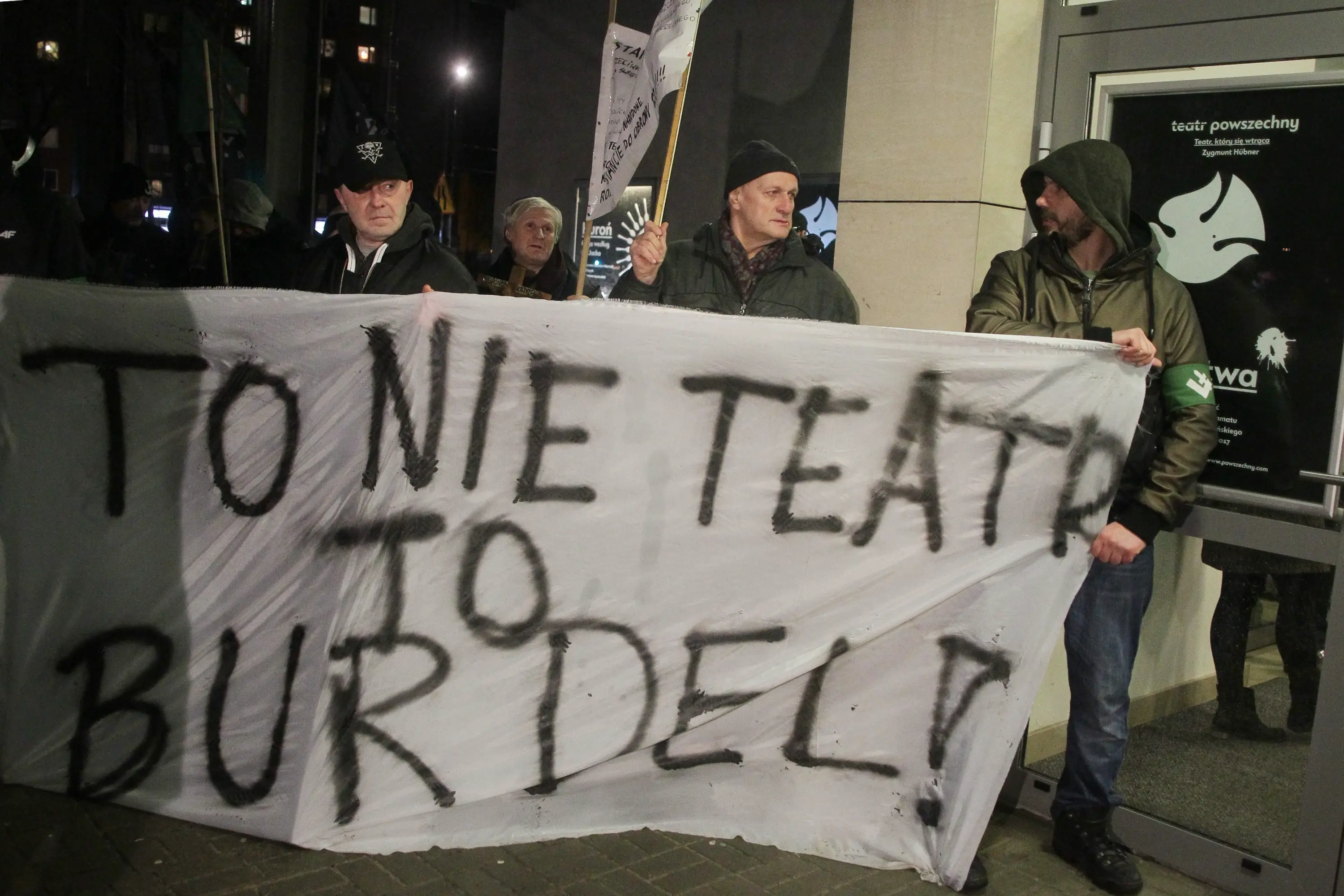 Po zmroku. Grupa protestujących przed Teatrem Powszechnym. Mężczyźni w kurtkach i czapkach trzymają biały materiał z niechlujnym napisem „To nie teatr, to burdel!”.